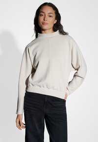 Maglione crema chiaro con colletto alto, finitura testurizzata e orlo elastico arricciato, abbinato a pantaloni in denim scuro. Design a vestibilità rilassata.