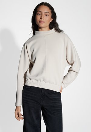Maglione crema chiaro con colletto alto, finitura testurizzata e orlo elastico arricciato, abbinato a pantaloni in denim scuro. Design a vestibilità rilassata.
