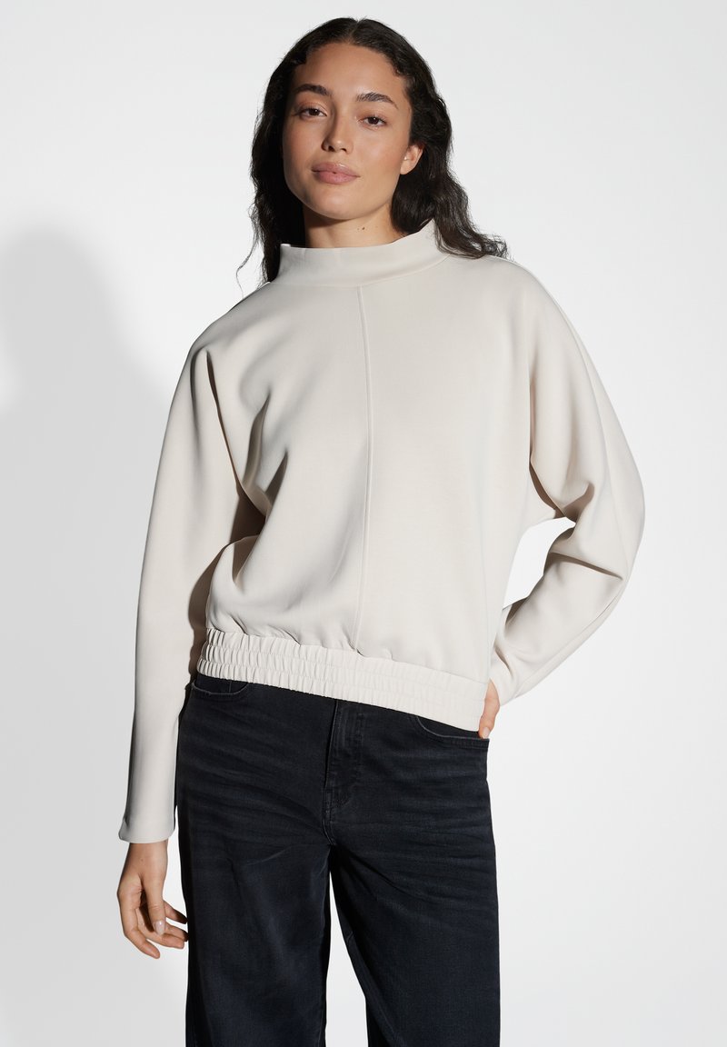 Maglione crema chiaro con colletto alto, finitura testurizzata e orlo elastico arricciato, abbinato a pantaloni in denim scuro. Design a vestibilità rilassata.
