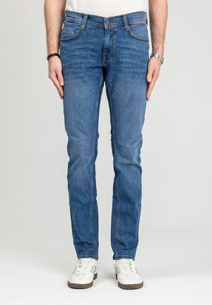 Mężczyzna ubrany w dopasowane niebieskie dżinsy, białe trampki, białą koszulę, zegarek na lewej ręce i bransoletkę na prawej, stoi na jednolitym tle.