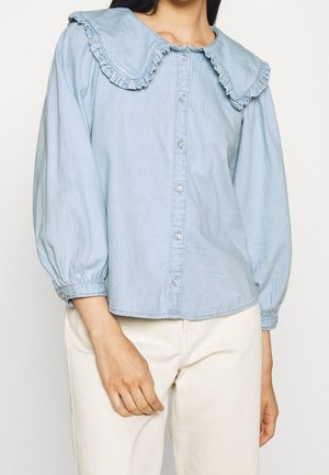 Blouse boutonnée bleu clair avec grand col à volants et manches trois-quarts bouffantes, associée à un pantalon blanc cassé.