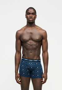 Μυώδης άνδρας στέκεται με το πρόσωπο προς τα εμπρός φορώντας μπλε μποξεράκια με επαναλαμβανόμενο μοτίβο λογότυπου "HUGO" σε απλό λευκό φόντο.