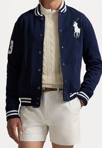 Mörkblå varsityjacka med vitrandig ribbad krage, logotyp, siffror och figurnära passform. Lager på en krämfärgad kabelstickad tröja.