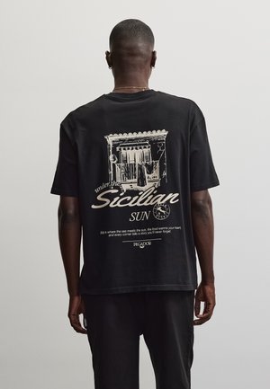 Man wearing black T-shirt with beige illustration of balcony and text "under the Sicilian sun" and smaller text about Sicily, standing against plain background.