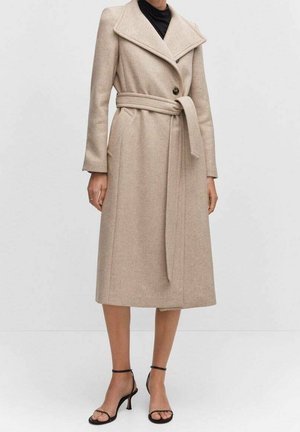 Femme portant un manteau en laine beige ceinturé avec de larges revers, sur un haut noir, accompagné de sandales à talons hauts noires à brides.