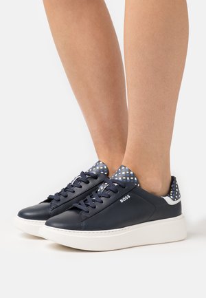 Námořnicky modré tenisky na šněrování s bílými platformními podrážkami, modro-bílým vzorovaným jazykem a detaily na patě, nošené na bosých nohou.