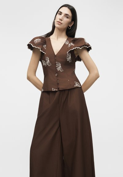 Γυναίκα που φοράει καφέ παντελόνι με φαρδιά πόδια και ένα καφέ μπλούζα κεντημένη με λουλούδια, με κυματιστά, φουσκωτά μανίκια.