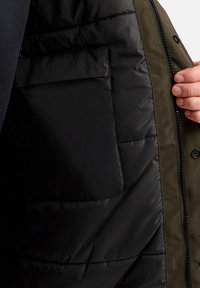 Giacca verde oliva con fodera in nylon nera, caratterizzata da una texture trapuntata. Include una zip visibile e una tasca a patta nera all'interno.