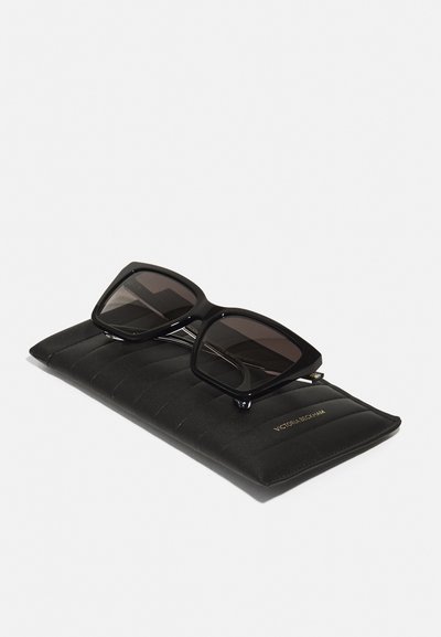 Black oversized square sunglasses with dark lenses, placed on a textured black case featuring a raised pattern and gold logo accent.