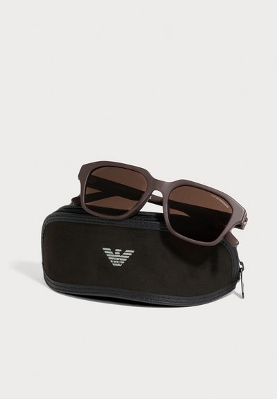 Des lunettes de soleil rectangulaires marron aux verres foncés reposent sur un étui noir zippé orné d’un logo blanc.