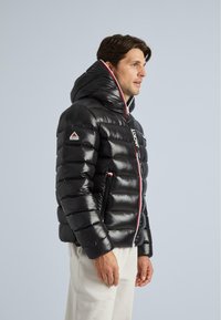 Jachetă puffer neagră cu guler înalt și glugă, având un material lucios, cusături orizontale și un fermoar contrastant cu accente roșii.