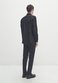 Maglietta nera a maniche lunghe con taglio dritto, dettaglio del giromanica sul retro, abbinata a pantaloni scuri e scarpe nere, su sfondo bianco.