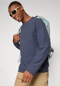 Marinefleece sweatshirt med rund hals, parret med en lyseblå rygsæk. Modellen bærer beige bukser og rammede solbriller.