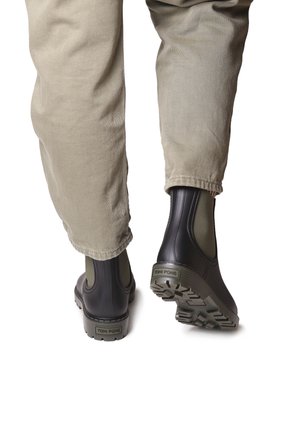 Botas de tobillo de goma negra con paneles laterales de elasticidad verde, suela texturizada y logotipo en el talón, combinadas con pantalones beige claros.