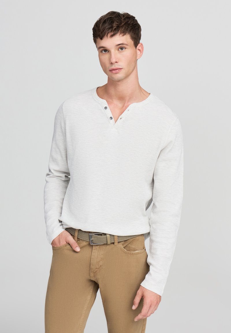 Maglietta a maniche lunghe henley grigia chiara con un motivo testurizzato, tre bottoni sul collo, abbinata a pantaloni color sabbia e una cintura intrecciata.