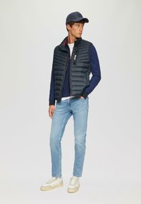 Marineblauer, gestepptes Weste über einem blauen, strukturierten Pullover, hellblaue Jeans und beiges Sneakers mit Akzenten. Abgerundet mit einer marineblauen Mütze.