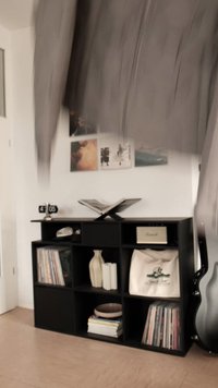 Giacca grigia in movimento contro un muro bianco. Un'unità di scaffalatura nera espone dischi in vinile, un vaso decorativo e una chitarra.