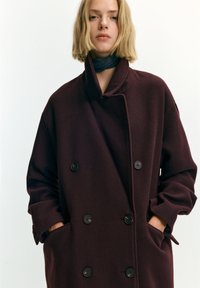 Blonde vrouw met een donker bordeauxrode dubbele rij knopen jas, handen in de zakken en een donkere nekkraag tegen een effen achtergrond.