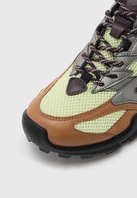 Világoszöld hálós felsőrészű cipő, barna bőr részletekkel, szürke ráhajtásokkal, fekete texturált talppal és fehér pettyes sötétbarna fűzőkkel.