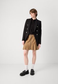 Schwarze zweireihige Blazer mit silbernen Knöpfen, kombiniert mit einem braunen plissierten Rock mit einem Reißverschluss an der Vorderseite und dekorativen Taschen sowie schwarzen Schuhen.