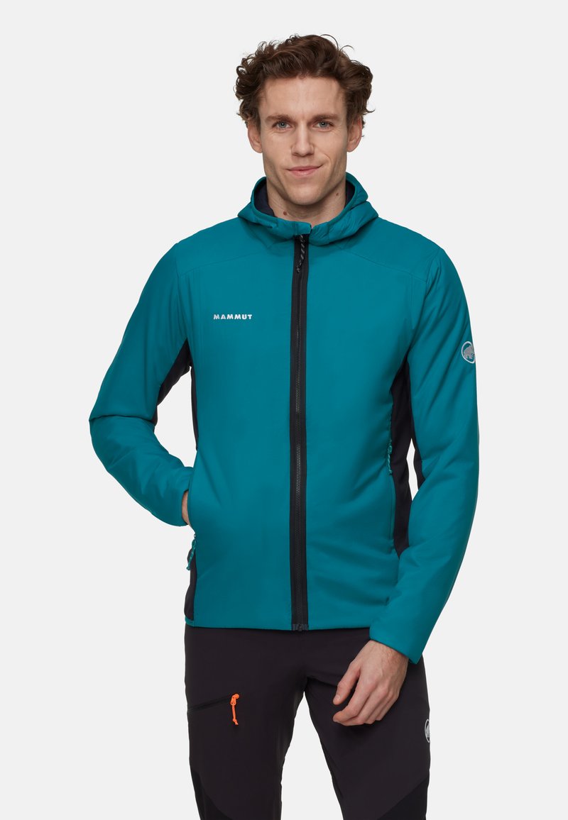 Homme portant une veste à capuche teal avec une fermeture éclair noire et un pantalon noir avec des détails de fermeture éclair orange, se tenant devant un fond uni clair.