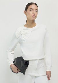 Frau in einem weißen Oberteil mit großem Schulter-Bow-Detail und weißen Hosen, die eine große schwarze Clutch-Tasche vor einem schlichten hellen Hintergrund hält.