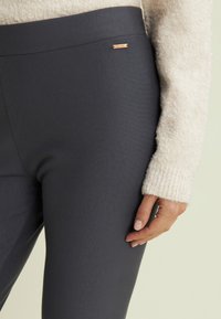 Pantaloni elasticizzati grigi con una texture liscia, caratterizzati da un'alta vita e un piccolo logo metallico sul lato. Maglione crema accogliente parzialmente visibile.