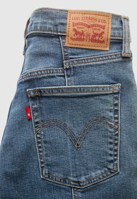Zone de la poche arrière d'un jean Levi's bleu montrant un patch de marque en cuir marron et une étiquette logo en tissu rouge sur un tissu denim avec des coutures visibles.