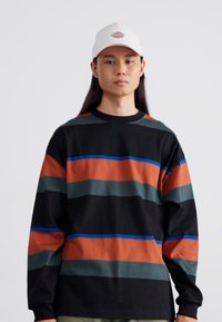 Camisa negra de manga larga con rayas horizontales en naranja, verde y azul. El modelo lleva una gorra blanca con un logo.