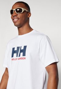 White short-sleeve t-shirt with large blue and red "HH" logo and "HELLY HANSEN" text. Sunglasses with a light frame and dark lenses.