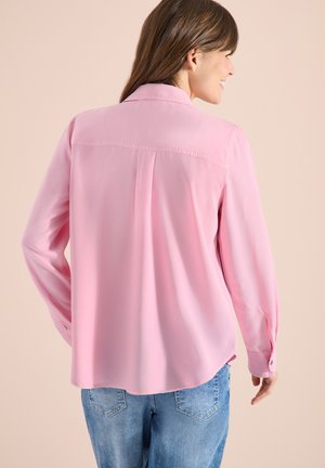 Femme aux cheveux longs bruns portant une chemise rose à manches longues ample et un jean bleu, de dos, souriant sur le côté.