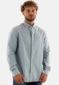 Camisa azul claro de tela suave con cuello, botones blancos y un pequeño logo en el pecho. Mangas largas incluidas.