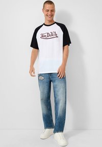 Bijela majica kratkih rukava s crnim raglan rukavima s crvenim "Von Dutch" logotipom, u kombinaciji s izblijedjelim plavim trapericama i bijelim tenisicama.