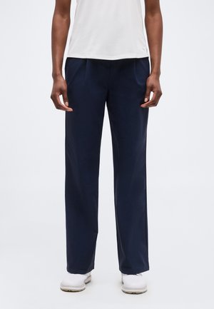 Personne portant un pantalon bleu marine droit, un haut blanc sans manches et des chaussures blanches, debout devant un fond uni.