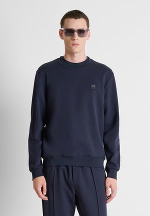 Hombre con sudadera azul marino oscuro y pantalones a juego con gafas de sol negras, de pie contra un fondo liso y claro.