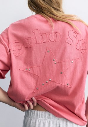 Personne portant un t-shirt corail avec le texte "Soho St." et une grande étoile contourée avec des clous en métal dans le dos, tenant l'ourlet du t-shirt.