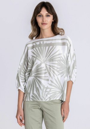 Vrouw draagt een losse witte blouse met een grijs abstract palmbladprint en driekwart mouwen, gecombineerd met lichtgroene broek.