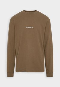 Ruskea pitkähiha-puuvillapaita, jossa on pyöreä kaula-aukko ja pieni valkoinen "DCSHOECO"-logo rinnassa. Sileä pinta.