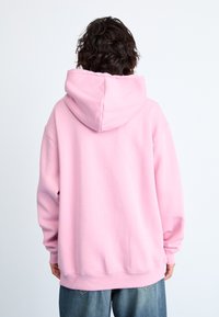 Personne avec des cheveux bouclés et foncés de dos, portant un sweat à capuche rose ample et un jean bleu, sur un fond clair uni.