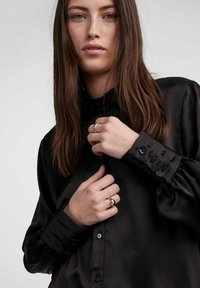 Black satin blouse with a high collar and long puffy sleeves. Features buttoned cuffs and a smooth, shiny texture. Model poses casually.