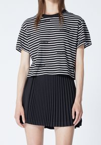 Randig svart och vit t-shirt med korta ärmar och rund halsringning, tillsammans med en svart veckad kjol. T-shirten har logotypdetaljer.
