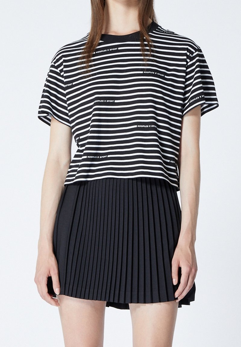 Randig svart och vit t-shirt med korta ärmar och rund halsringning, tillsammans med en svart veckad kjol. T-shirten har logotypdetaljer.