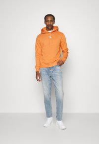 Felpa arancione con cappuccio, stringhe e logo, abbinata a jeans azzurri chiari e sneaker bianche. Design semplice e casual, con vestibilità confortevole.
