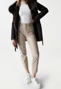 Mujer vistiendo pantalones cortos ajustados beige, zapatillas blancas, camiseta blanca y chaqueta de piel sintética marrón oscuro en un fondo blanco.