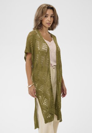 Kvinde iført lyserød blonderbluse, beige bukser og olivengrøn åbenstrikket lang cardigan med korte ærmer, poserer mod ensfarvet baggrund.