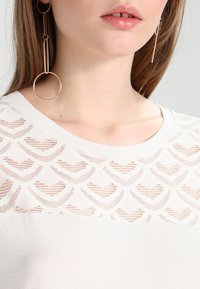 Maglietta bianca a maglia con un motivo a cuore nella parte superiore in rete, sezione inferiore liscia e scollatura rotonda. Orecchini lunghi con design circolare.