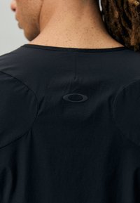 Črna majica s kratkimi rokavi z gladko, mat površino, okroglim ovratnikom in okroglimi oblikovalskimi dodatki na ramah. Logo je prikazan osrednjo na hrbtni strani.