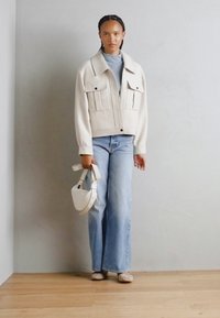 Kremfarget cropped jakke med to foran lommer, kombinert med lyseblå vide jeans og en liten hvit håndveske. Enkle teksturerte sko.