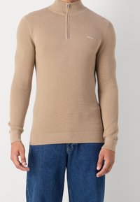 Pullover beige in maglia con texture e colletto a zip quarti, indossato con jeans blu a vestibilità ampia, mostrato dalla nuca ai fianchi.