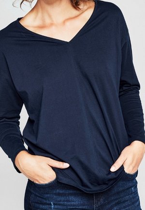 Woman wearing a loose-fitting navy blue long-sleeve V-neck shirt with hands partially in dark blue jeans pockets.